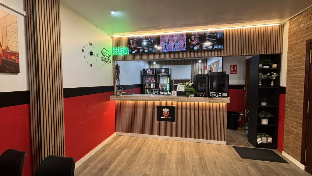 Mostrador de atención al cliente de La Casa de Tacos, decorado con listones de madera iluminados. Arriba se ven tres pantallas digitales con el menú del restaurante y un cartel de neón verde que indica "Pide Aquí".