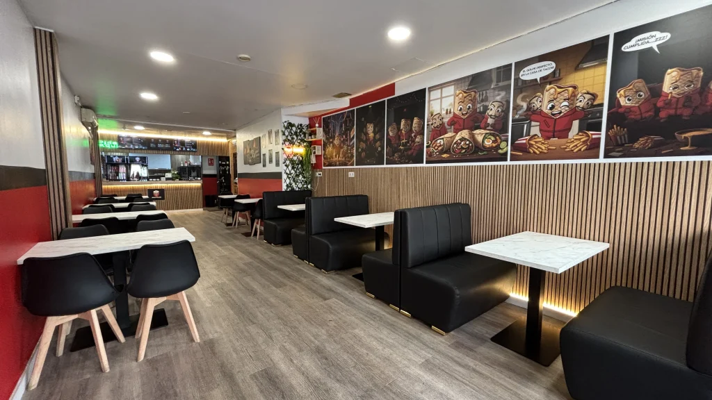 Interior del restaurante La Casa de Tacos, mostrando mesas blancas con sofás negros acolchados. En la pared de madera destaca un gran mural decorativo tipo cómic protagonizado por burritos y la máscara de Dalí de La Casa de Papel.